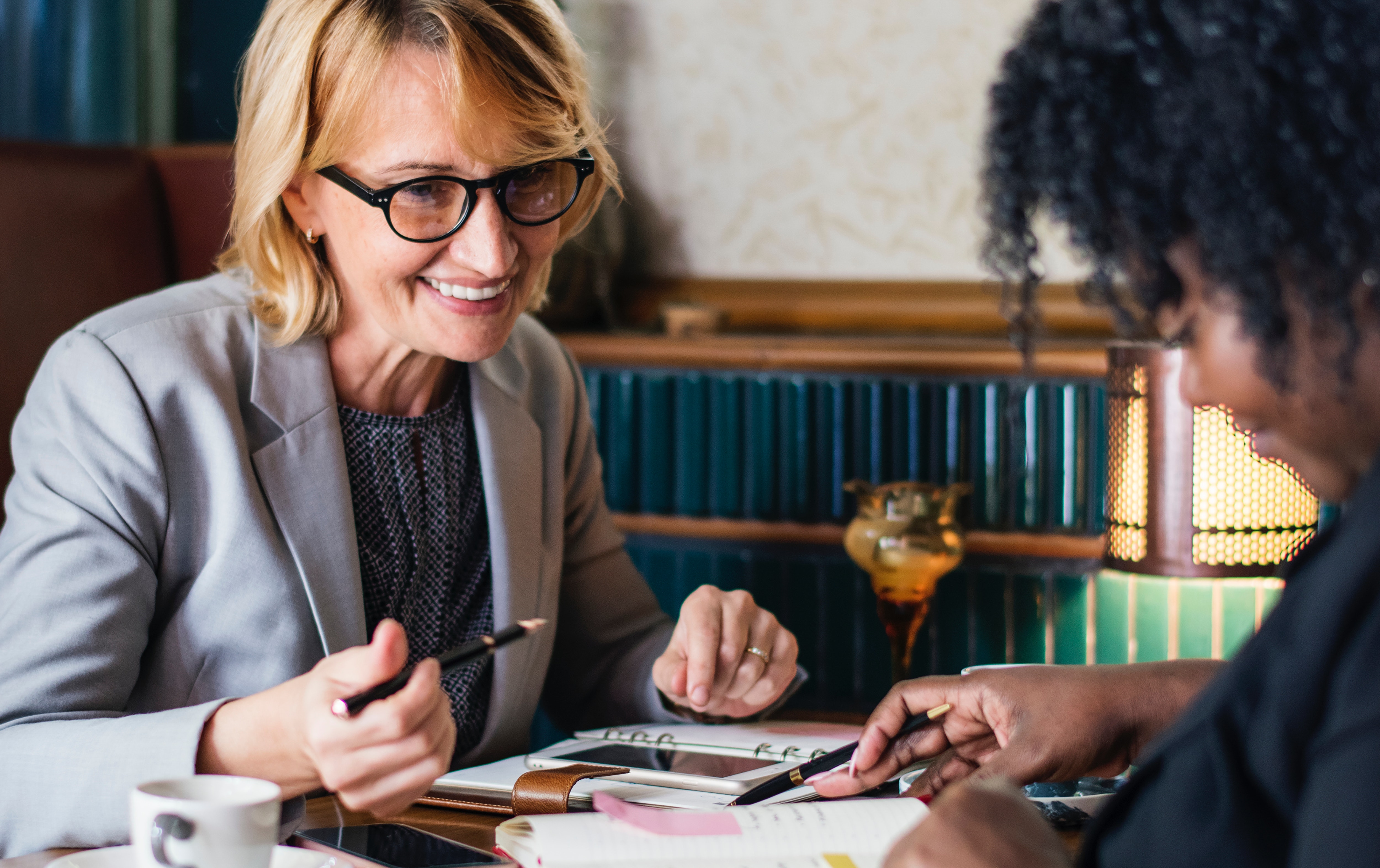 A woman in glasses and a gray blazer smiles while discussing paperwork with another person at a table, conveying a collaborative and friendly atmosphere.