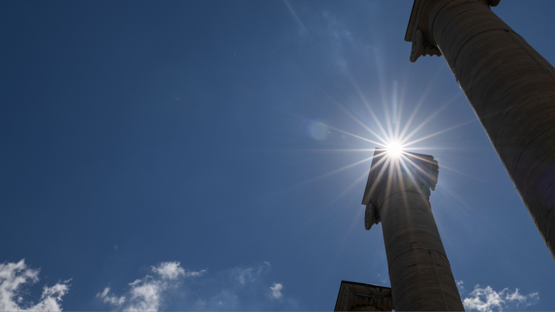 Sunlight peeks between two ancient columns against a clear blue sky, casting dramatic starburst rays, evoking a sense of history and wonder.