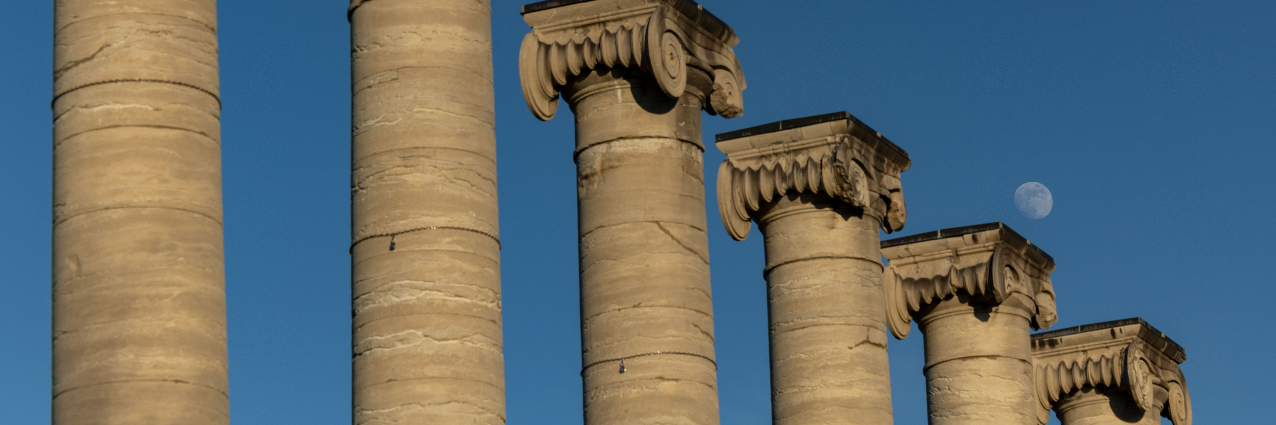 Row of five stone columns with ornate capitals, set against a clear blue sky. A small white moon is visible, adding a sense of tranquility.