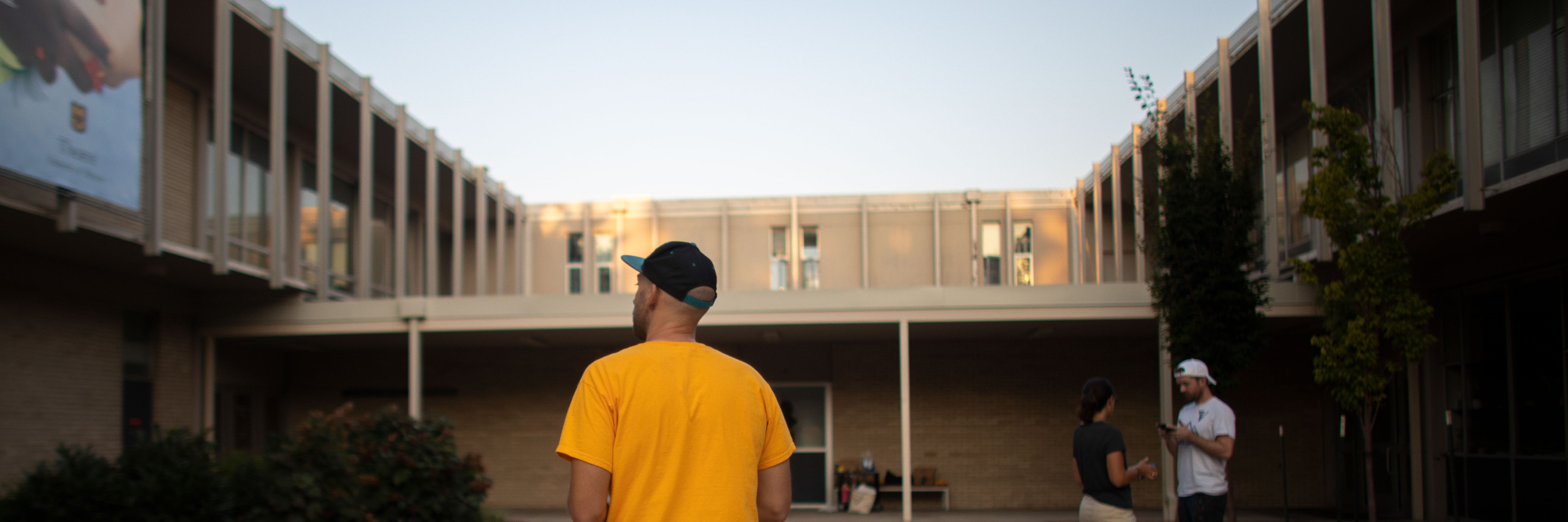 A person in a yellow shirt and cap stands in a courtyard, framed by modern buildings. Two others converse nearby, under a clear sky at dusk.