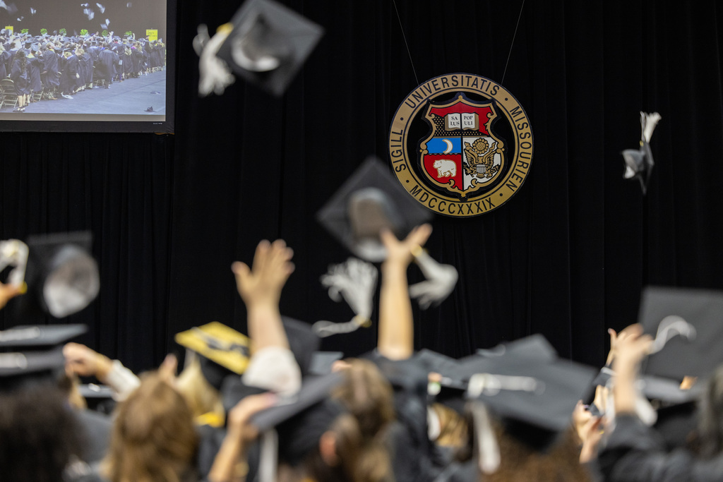 Graduates in caps and gowns cheer, tossing caps into the air against a backdrop featuring a university seal, capturing the joy of commencement.