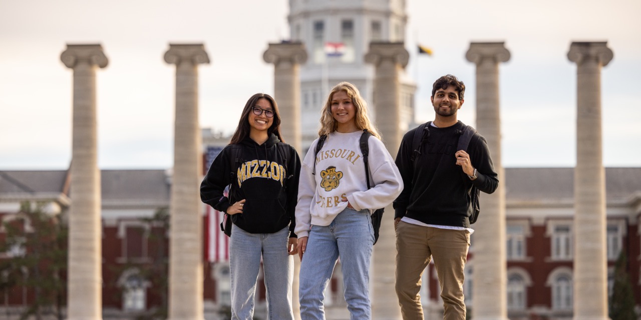 Three students stand in front of university columns, wearing casual clothes and backpacks. They're smiling, embodying a cheerful and academic atmosphere.