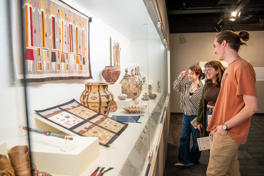 Three people admire traditional textiles and pottery in a well-lit museum display. They appear engaged and curious.
