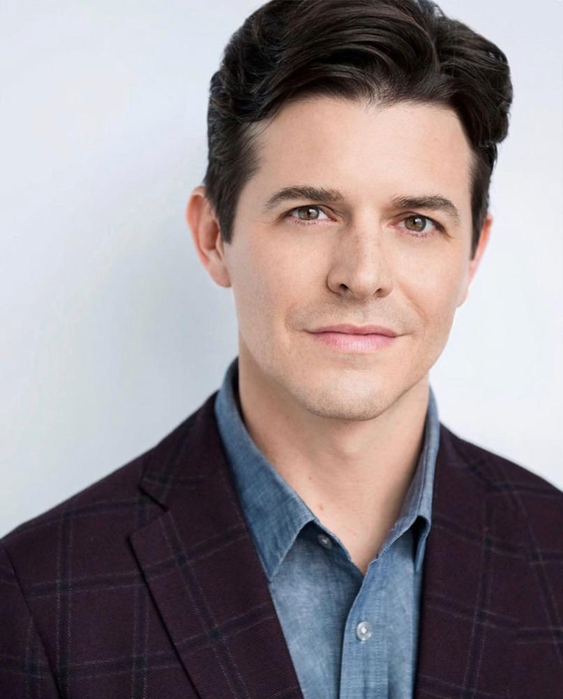 head-and-shoulders portrait of Jason Forbach wearing a blue shirt and dark plaid blazer against a light gray background.