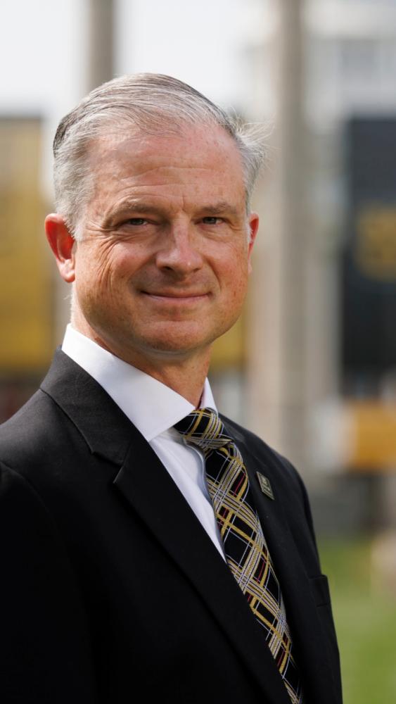 Dean Drury wearing a black suit with a plaid tie stands outdoors, smiling softly. The background is blurred, suggesting an urban setting.