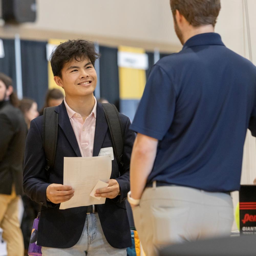 A young man in a blazer and pink shirt smiles while holding papers, engaging with another person in a blue shirt at an event, conveying friendliness.
