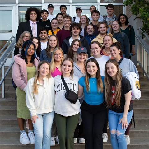 A&S Student Council stands on outdoor stairs, smiling at the camera. They are casually dressed, conveying camaraderie and a joyful atmosphere.