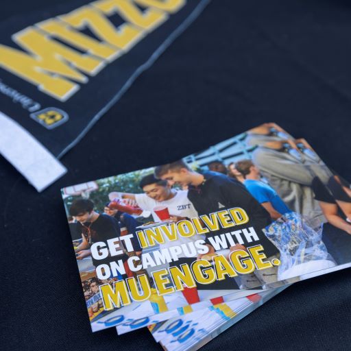 Students engaging at campus event, smiling and interacting. A flyer with the text "Get involved on campus with MU Engage," exudes an inviting tone.