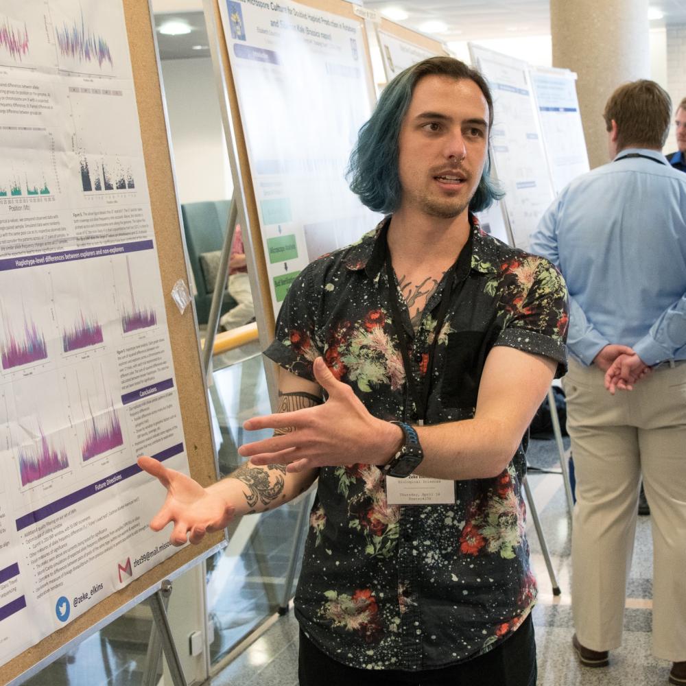 A person with blue hair presents a scientific poster at a conference. They wear a patterned shirt and gesture expressively, conveying enthusiasm.