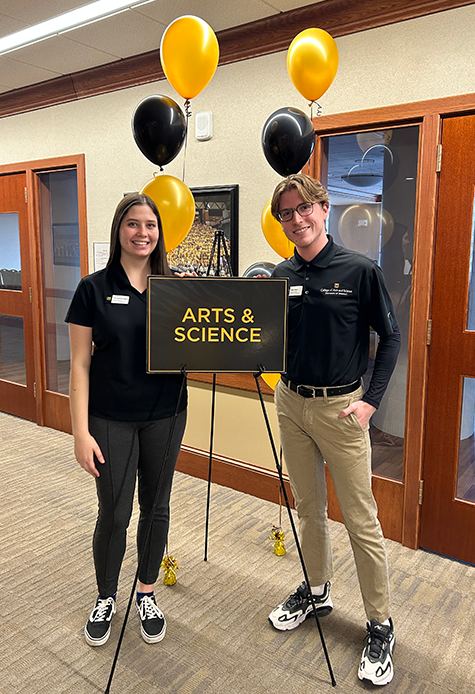 Two ambassadors stand smiling beside a sign reading "Arts & Science," with black and yellow balloons in the background. The setting is bright and welcoming.