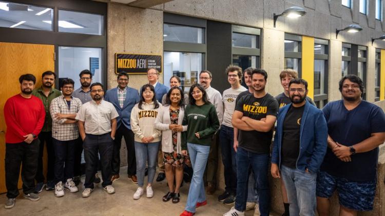 A group of people stand and pose for a photo in a hallway beside a sign that reads "Mizzou AERI," with doors and windows visible in the background.