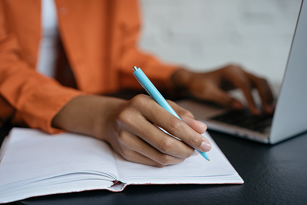 A person in an orange shirt writes in a notebook with a blue pen while typing on a laptop. The setting suggests focused multitasking and productivity.