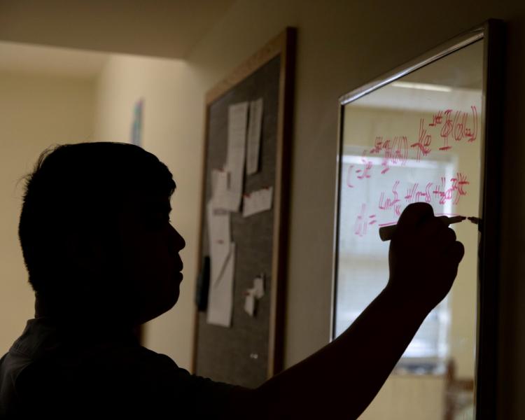 A person writes equations on a clear board with red marker. The setting is a dimly lit room with a bulletin board in the background.