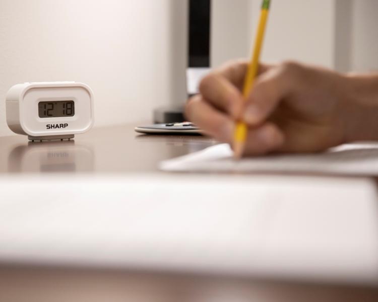 A close-up image shows a person writing with a pencil on a desk. A white digital clock displays 12:18. The scene conveys focus and concentration.