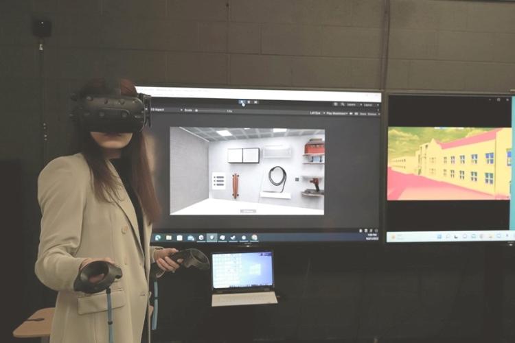 A person in a beige blazer uses VR gear, holding controllers, in front of screens displaying virtual environments, showing modern and colorful scenes.