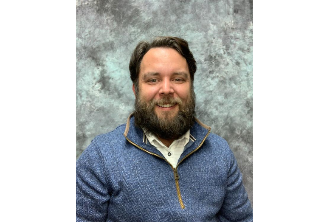 James Kendall in a blue sweater smiles warmly against a textured gray backdrop, conveying a friendly and approachable demeanor.