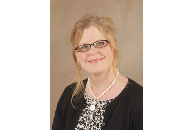 Kirsten Pape with glasses and a friendly smile, wearing a black and white floral blouse, black cardigan, pearl necklace, and earrings, against a neutral backdrop.