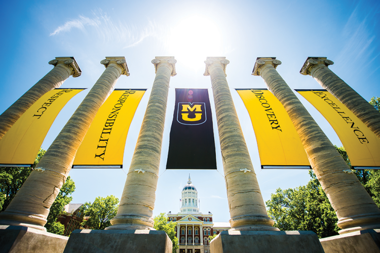 Historic stone columns under a bright sky display large yellow banners with the University of Missouri logo. A grand building stands in the background.