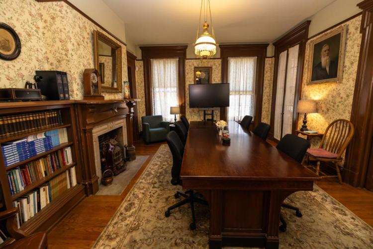 A vintage-style office with a wooden table, six chairs, a computer, a bookshelf, and a fireplace. Warm lighting and floral wallpaper create a cozy ambiance.