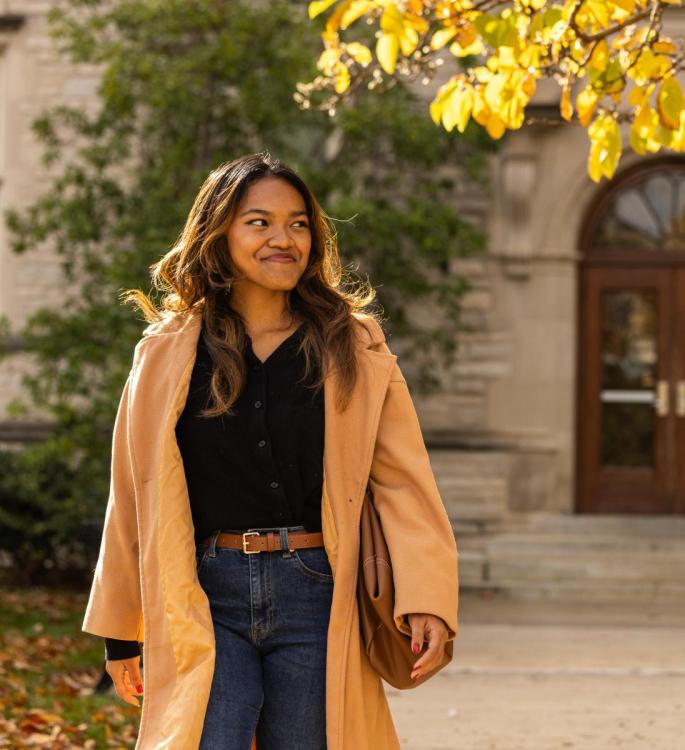 Woman smiling, wearing a beige coat and jeans, walks outside in autumn sun with golden leaves overhead. She seems content and relaxed.